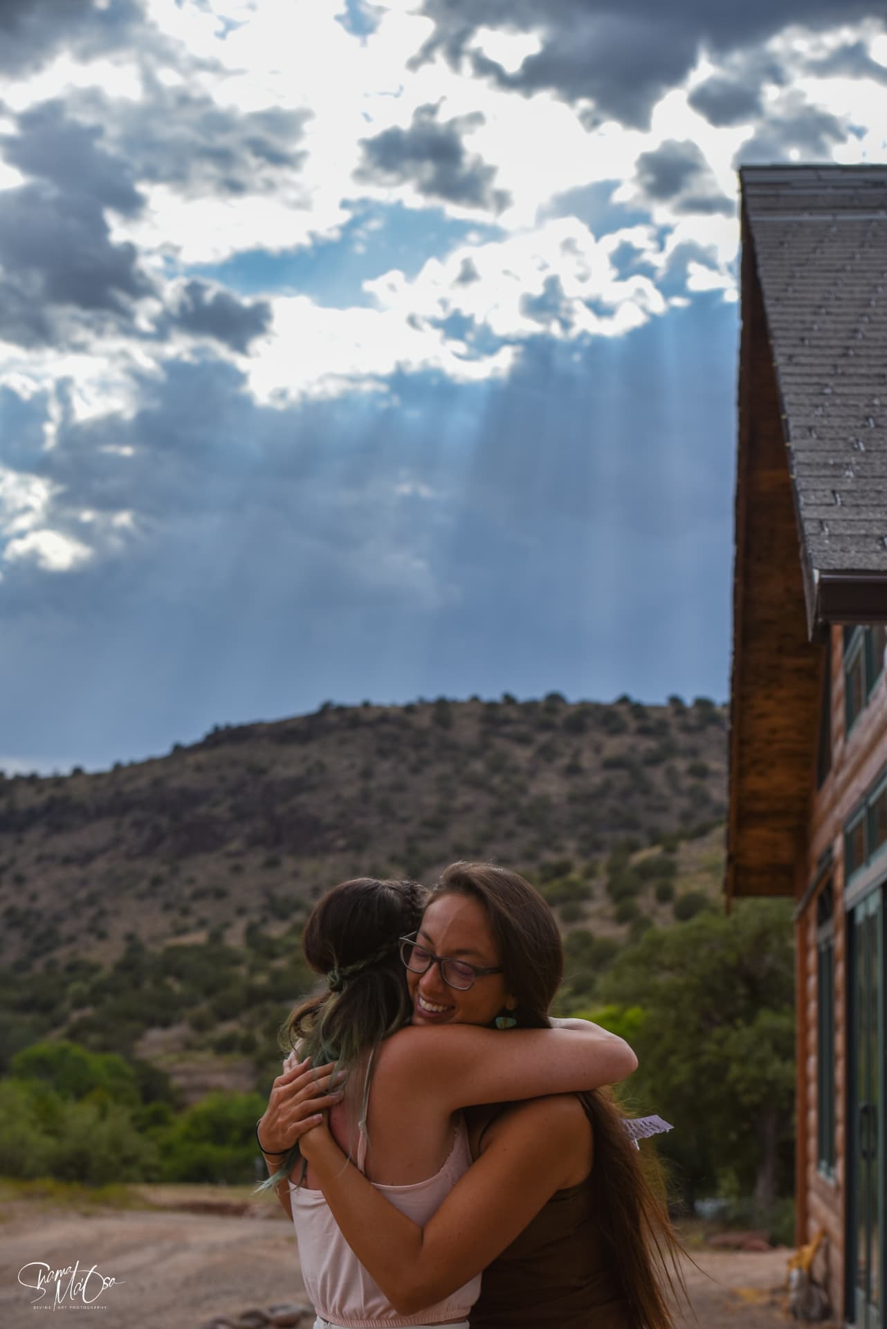 Two women holding each other in a long embrace beneath a cloud-filled mountain sky
